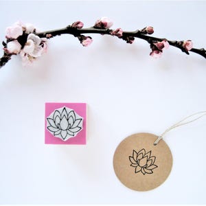 May include: A square rubber stamp with a pink base featuring a black line art lotus flower design, positioned next to a round brown paper tag stamped with the same lotus image, all displayed below a dark branch with small light pink blossoms.