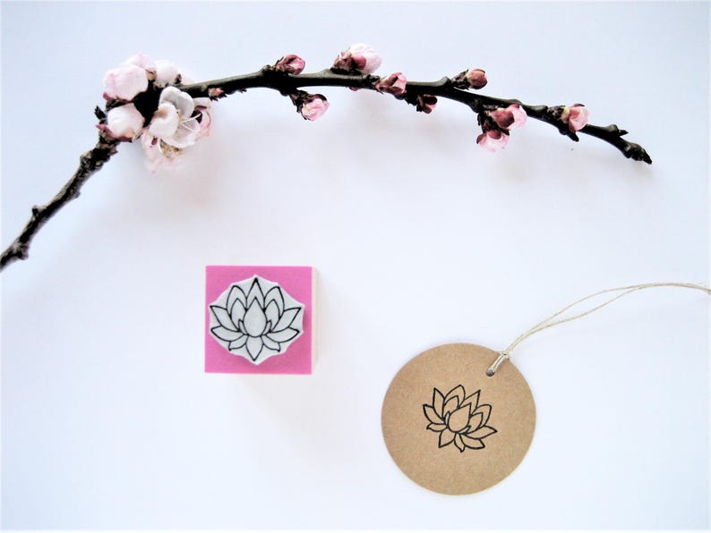 May include: A square rubber stamp with a pink base featuring a black line art lotus flower design, positioned next to a round brown paper tag stamped with the same lotus image, all displayed below a dark branch with small light pink blossoms.