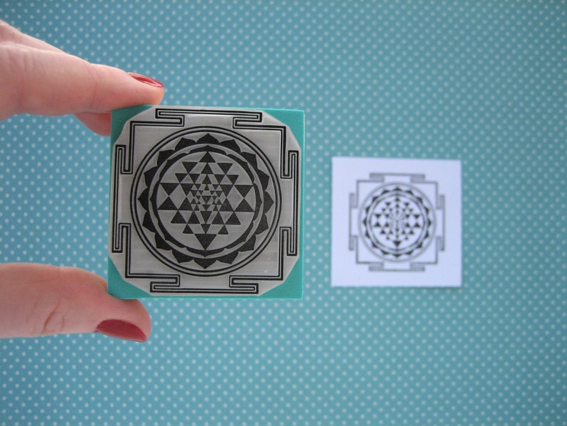 May include: A square stamp with a geometric design, held by a hand. The stamp has a teal base and a silver surface with a black, intricate pattern. A printed version of the design is next to the stamp. The background is light blue with white polka dots.