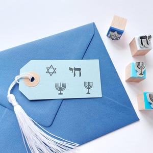 May include: A light blue paper gift tag with a white tassel, featuring black stamped designs of a Star of David, the Hebrew word Chai, and two menorahs, resting on a blue envelope next to four small wooden stamps with blue bases.