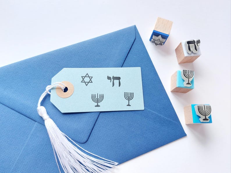 May include: A light blue paper gift tag with a white tassel, featuring black stamped designs of a Star of David, the Hebrew word Chai, and two menorahs, resting on a blue envelope next to four small wooden stamps with blue bases.