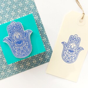 May include: A blue rubber stamp featuring a detailed Hamsa hand design with an eye motif, mounted on a square teal block. Beside it, a cream-colored paper gift tag displays a crisp blue ink impression of the same Hamsa hand pattern.