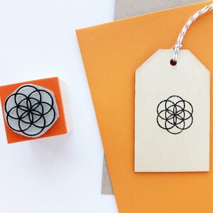Op de afbeelding: Een houten stempel met een geometrisch ontwerp in zwart en wit. De stempel bevindt zich op een fel oranje oppervlak. Een houten label met hetzelfde ontwerp bevindt zich op een fel oranje oppervlak.