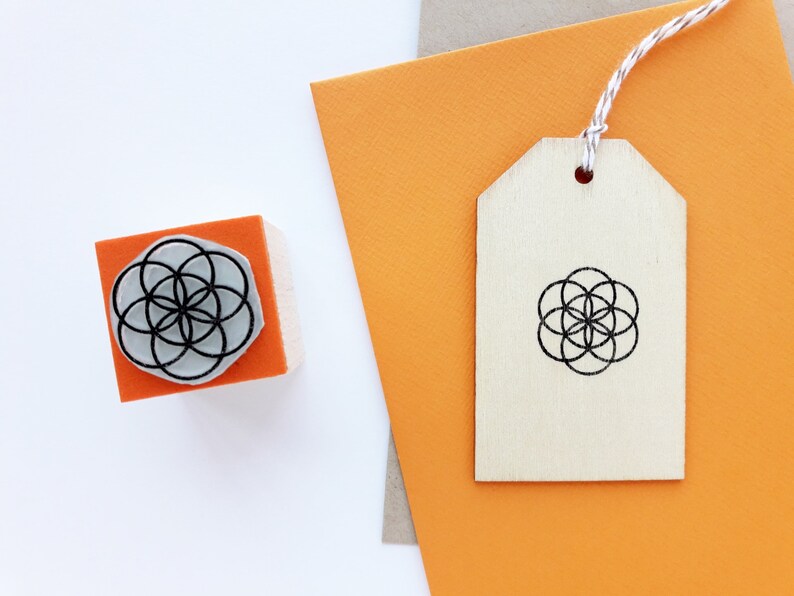 Op de afbeelding: Een houten stempel met een geometrisch ontwerp in zwart en wit. De stempel bevindt zich op een fel oranje oppervlak. Een houten label met hetzelfde ontwerp bevindt zich op een fel oranje oppervlak.