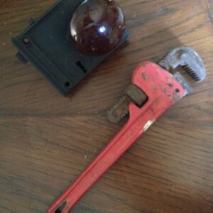 May include: A red adjustable pipe wrench and a black door lock with a brown knob are displayed on a wooden surface. The wrench shows signs of wear, and the lock has a keyhole.