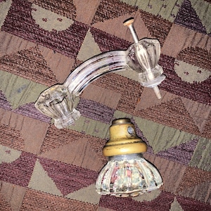 May include: A clear glass cabinet handle with a curved design and a faceted knob. The handle is attached to a metal rod with a nut and washer. A matching glass knob with a gold-colored metal base is also present.