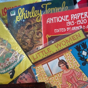 May include: A collection of vintage paper doll books featuring a variety of themes, including Shirley Temple, Little Women, and Antique Paper Dolls. The books are in various colors, including red, yellow, blue, and green. The covers feature illustrations of dolls and text.