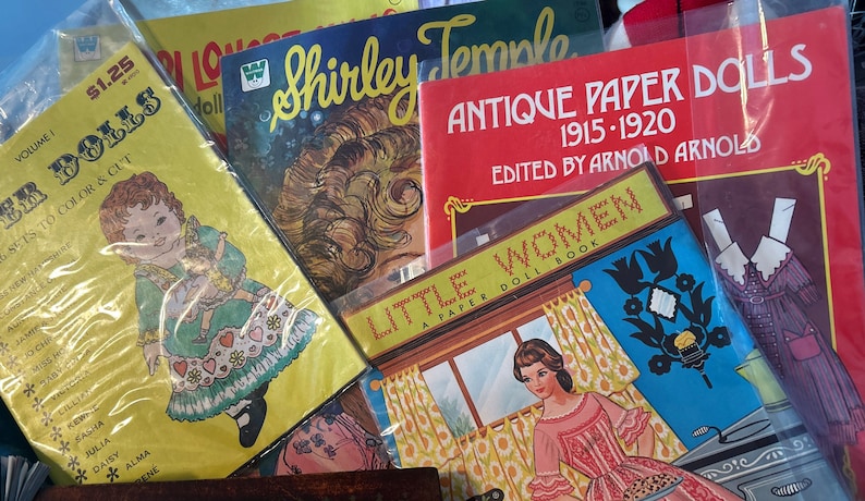 May include: A collection of vintage paper doll books featuring a variety of themes, including Shirley Temple, Little Women, and Antique Paper Dolls. The books are in various colors, including red, yellow, blue, and green. The covers feature illustrations of dolls and text.