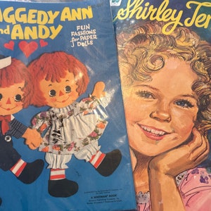 May include: Two paper doll books, one featuring Raggedy Ann and Andy with the text "Fun Fashions for Paper Dolls" and the other featuring Shirley Temple with the text "Paper Doll Book".