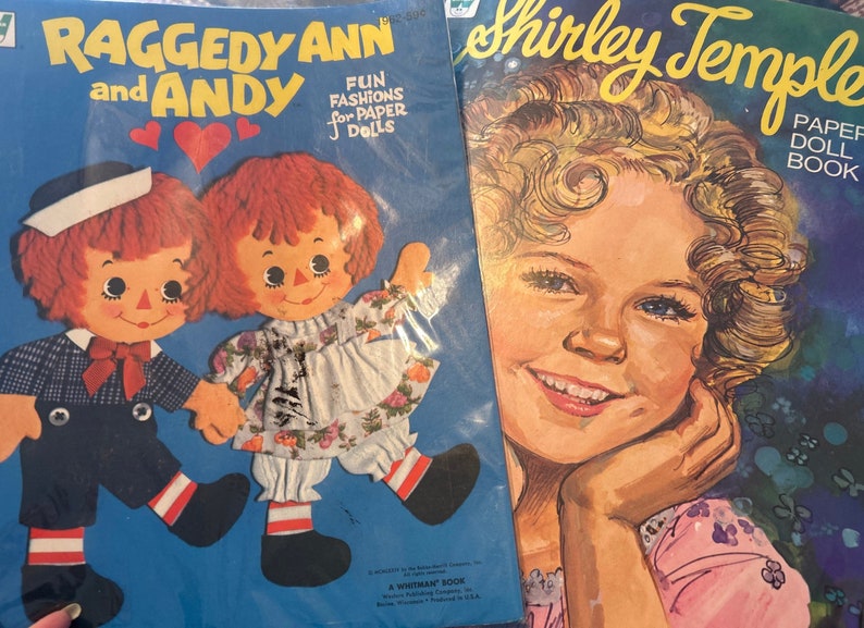 May include: Two paper doll books, one featuring Raggedy Ann and Andy with the text "Fun Fashions for Paper Dolls" and the other featuring Shirley Temple with the text "Paper Doll Book".