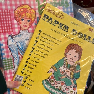 May include: A yellow paper doll book with the title "Stitch 'n Sew Paper Dolls" and the text "16 Sets to Color & Cut". The book features a colorful illustration of a girl in a green dress holding a smaller doll. The book cover also includes a list of doll names, including "Miss New Hampshire Constance of 1860", "Auntie Nellie", "Jamie & Jennie", and "Jo Christina".