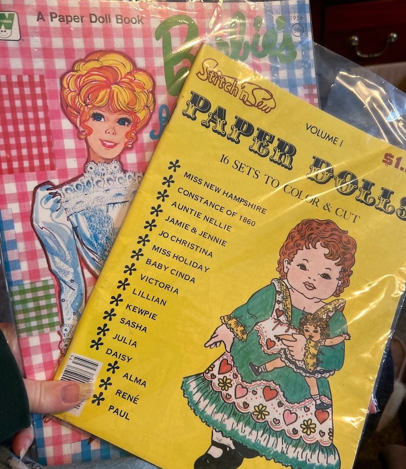 May include: A yellow paper doll book with the title "Stitch 'n Sew Paper Dolls" and the text "16 Sets to Color & Cut". The book features a colorful illustration of a girl in a green dress holding a smaller doll. The book cover also includes a list of doll names, including "Miss New Hampshire Constance of 1860", "Auntie Nellie", "Jamie & Jennie", and "Jo Christina".