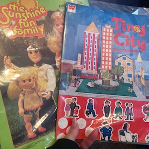 May include: A vintage press-out book titled "Tiny City" with colorful illustrations of buildings, trees, and people. The book is open to a page with a scene of a city park with a fountain and benches. The book is part of a set of vintage toys and games.
