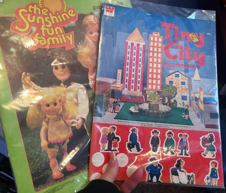 May include: A vintage press-out book titled "Tiny City" with colorful illustrations of buildings, trees, and people. The book is open to a page with a scene of a city park with a fountain and benches. The book is part of a set of vintage toys and games.