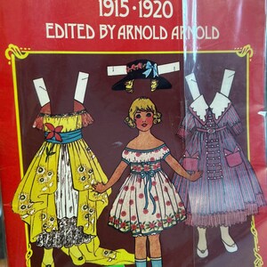 May include: Antique paper dolls from 1915-1920, edited by Arnold Arnold. The dolls are dressed in vintage clothing, including a yellow dress with a blue sash, a pink dress with a blue sash, and a purple dress with a white collar. The dolls are holding a hat, a purse, and a flower. The book is priced at $5.00.