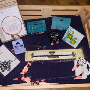 May include: A wooden crate filled with various jewelry items, including necklaces, bracelets, earrings, and a charm bracelet with a guardian angel charm. The jewelry is packaged on small cards with the brand names "Toni", "Belle", "daire's", and "funky pretty".