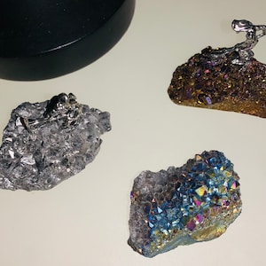 May include: Three different crystal clusters with silver metal figurines on top. The crystals are iridescent and have a variety of colors, including purple, blue, gold, and silver.