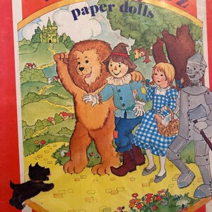 May include: A vintage Wizard of Oz paper doll book featuring a colorful illustration of Dorothy, the Scarecrow, the Tin Man, and the Cowardly Lion walking down the yellow brick road. The book cover includes the title "Wizard of Oz paper dolls" in purple and yellow lettering.