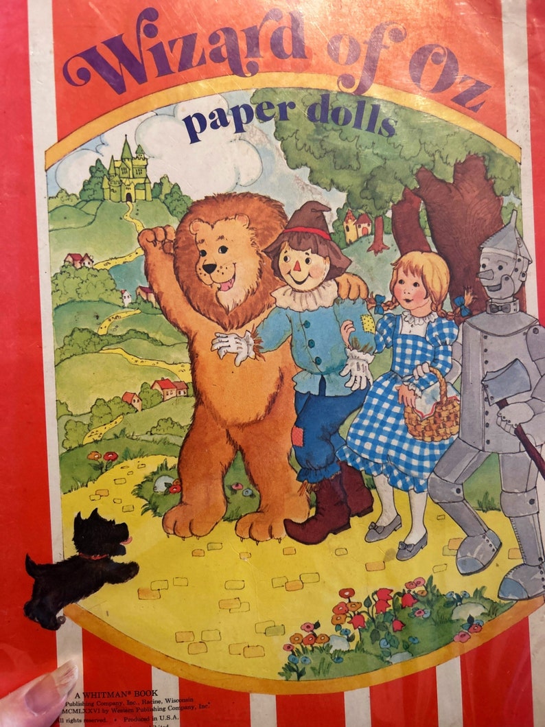 May include: A vintage Wizard of Oz paper doll book featuring a colorful illustration of Dorothy, the Scarecrow, the Tin Man, and the Cowardly Lion walking down the yellow brick road. The book cover includes the title "Wizard of Oz paper dolls" in purple and yellow lettering.