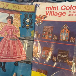May include: Two vintage paper doll books. The first book is titled "Little Women, A Paper Doll Book" and features a doll dressed in a pink dress holding a pie. The second book is titled "Mini Colonial Village to press out and assemble" and features illustrations of colonial houses.