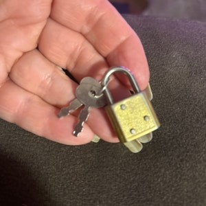 May include: A small, brass-colored padlock with a silver shackle and two matching keys. The padlock has a rectangular body with rounded corners and is secured with a key. The keys are silver-colored and have a simple design.