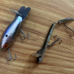 May include: Two fishing lures, one silver with a blue and orange design and one brown with a silver eye. Both lures have treble hooks.