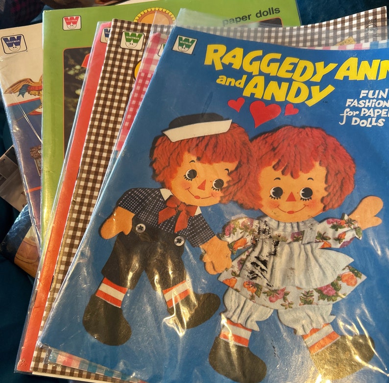May include: A stack of paper doll books featuring Raggedy Ann and Andy. The cover of the top book shows Raggedy Ann and Andy in colorful outfits on a blue background. The text on the cover reads "Raggedy Ann and Andy Fun Fashion for Paper Dolls".
