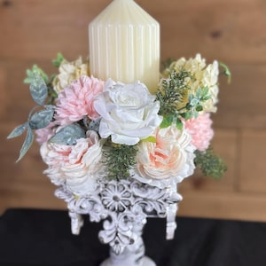 Könnte beinhalten: Eine cremefarbene Säulenkerze sitzt auf einem weißen, verzierten Kerzenhalter, umgeben von rosa, weißen und pfirsichfarbenen Kunstblumen und Grün. Der Kerzenhalter hat ein Used-Finish.