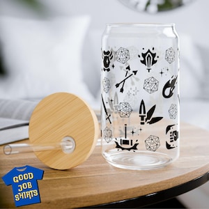 May include: A clear glass can-shaped drinking glass with black graphics of dice, swords, and other fantasy symbols. It has a bamboo lid and a clear straw. The text "GOOD JOB SHIRTS" is on a blue t-shirt graphic.