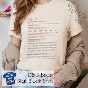 May include: Beige t-shirt with the text "D&D Bride Stat Block Shirt" and a graphic. The shirt features a stat block with game information, including stats like STR, DEX, and CHA, and details about the Bride's abilities. Two dice are visible in the background.