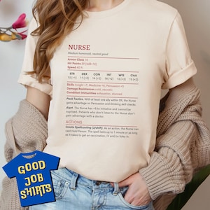 May include: A beige t-shirt with a graphic design of a character profile for a "NURSE" role-playing game. The character has a strength score of 13, dexterity of 11, constitution of 14, intelligence of 14, wisdom of 16, and charisma of 15. The text "GOOD JOB SHIRTS" is printed on the bottom of the shirt.