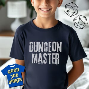 May include: A navy blue t-shirt with the words "DUNGEON MASTER" in distressed white lettering. The shirt is being worn by a person. Two black and white geometric dice are in the background. A small blue graphic with the words "GOOD JOB SHIRTS" is in the lower left corner.