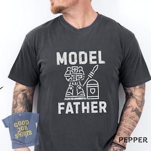 May include: A charcoal gray short-sleeve t-shirt featuring the white distressed text "MODEL FATHER" with a graphic of a miniature figure and a paintbrush. A smaller blue t-shirt with the yellow text "GOOD JOB SHIRTS" is visible in the corner.