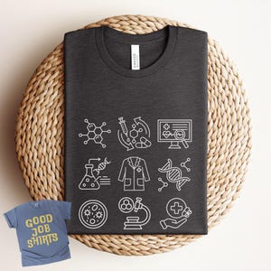 May include: Dark gray t-shirt with white science-themed graphics, including molecular structures, lab equipment, and medical symbols. A smaller blue t-shirt with the text "GOOD JOB SHIRTS" is also visible.