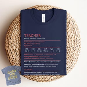 May include: Dark blue t-shirt with the word "TEACHER" in pink, featuring game stats. A smaller light blue t-shirt with "GOOD JOB SHIRTS" is also visible. The shirt is displayed on a woven, circular surface.