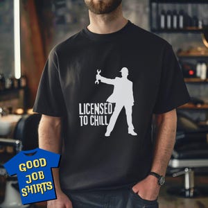 May include: Black t-shirt with a white graphic of a person holding a wrench, wearing a hard hat and suit. The text "LICENSED TO CHILL" is below the graphic. The shirt has short sleeves.