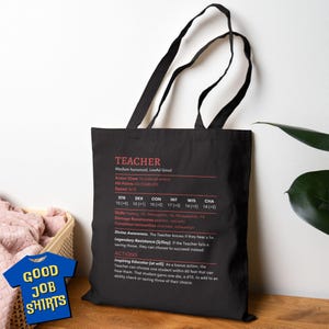 May include: Black tote bag with a white graphic of a teacher character sheet. The text on the graphic reads "TEACHER" and includes stats like "STR", "DEX", "CON", "INT", "WIS", "CHA", "Armor Class", "Hit Points", "Speed", "Skills", "Damage Resistances", "Condition Immunities", "Divine Awareness", "Legendary Resistance", and "Actions".