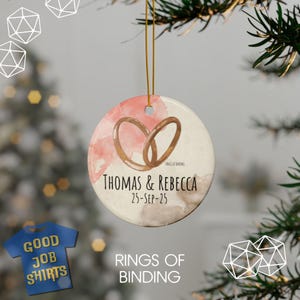 May include: Round ceramic ornament with a watercolor design in pink and brown, featuring two intertwined rings. The ornament is personalized with the names "THOMAS & REBECCA" and the date "25-SEP-25". The text "RINGS OF BINDING" is also present.