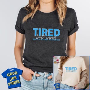 May include: A dark gray t-shirt with the text "TIRED but wired GRAVES DISEASE AWARENESS" in blue. The shirt is being modeled by a person wearing blue jeans.