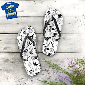 May include: A pair of white flip-flops with a black and white pattern of dice, swords, and other gaming-related icons. The flip-flops have black straps and are displayed on a light wood surface. A blue t-shirt with the text "GOOD JOB SHIRTS" is in the background.