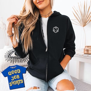 May include: Black zip-up hoodie with a white geometric design on the chest. The hoodie features a full zip, a hood, and pockets. The model is wearing a white t-shirt underneath. The image also includes a blue t-shirt with the text "GOOD JOB SHIRTS".