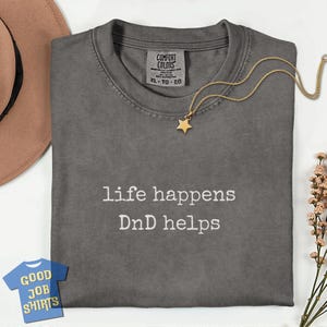 May include: A gray Comfort Colors t-shirt with the text "life happens DnD helps" in white. A gold star pendant necklace and a small blue and white "Good Job Shirts" tag are also visible.