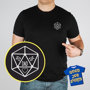 May include: Black t-shirt with a white graphic of a 20-sided die, the word "DAD", and a heart. A smaller die graphic is on the chest. The text "GOOD JOB SHIRTS" is on a blue t-shirt graphic.