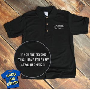 May include: Black polo shirt with the text "IF YOU ARE READING THIS, I HAVE FAILED MY STEALTH CHECK" printed on the front. The text is in white and includes a white 20-sided die icon.