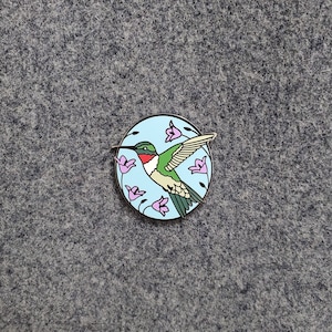 Puede incluir: Un colibrí verde y rojo con un pico largo vuela sobre un fondo azul con flores rosas. El colibrí es un pin de metal con un acabado brillante.