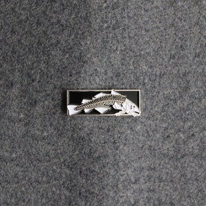 Puede incluir: Un pin de esmalte negro con un diseño de esqueleto de pez blanco. El pin tiene forma rectangular y tiene un acabado plateado.