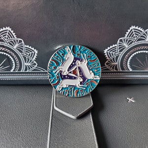 May include: A silver enamel pin with a circular design featuring three white rabbits hopping in a circle around a blue and purple triangle. The background is a teal blue with white stars and orange flowers.