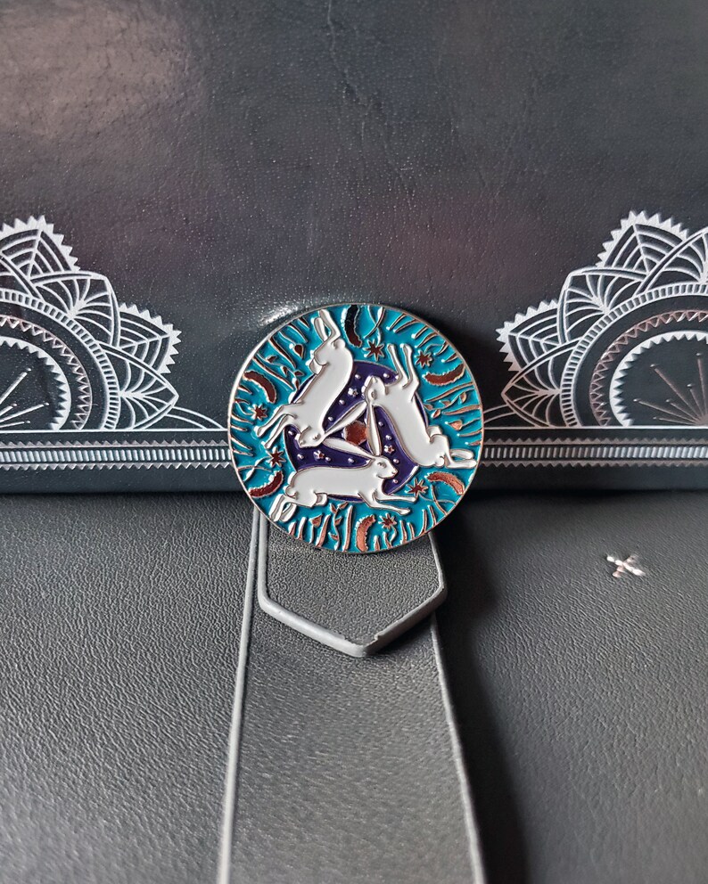 May include: A silver enamel pin with a circular design featuring three white rabbits hopping in a circle around a blue and purple triangle. The background is a teal blue with white stars and orange flowers.