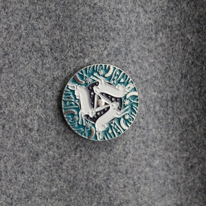 Puede incluir: Un pin de esmalte azul verdoso con borde plateado que presenta tres conejos blancos en un círculo. Los conejos están en posición de salto y están rodeados de un patrón de estrellas y hojas.