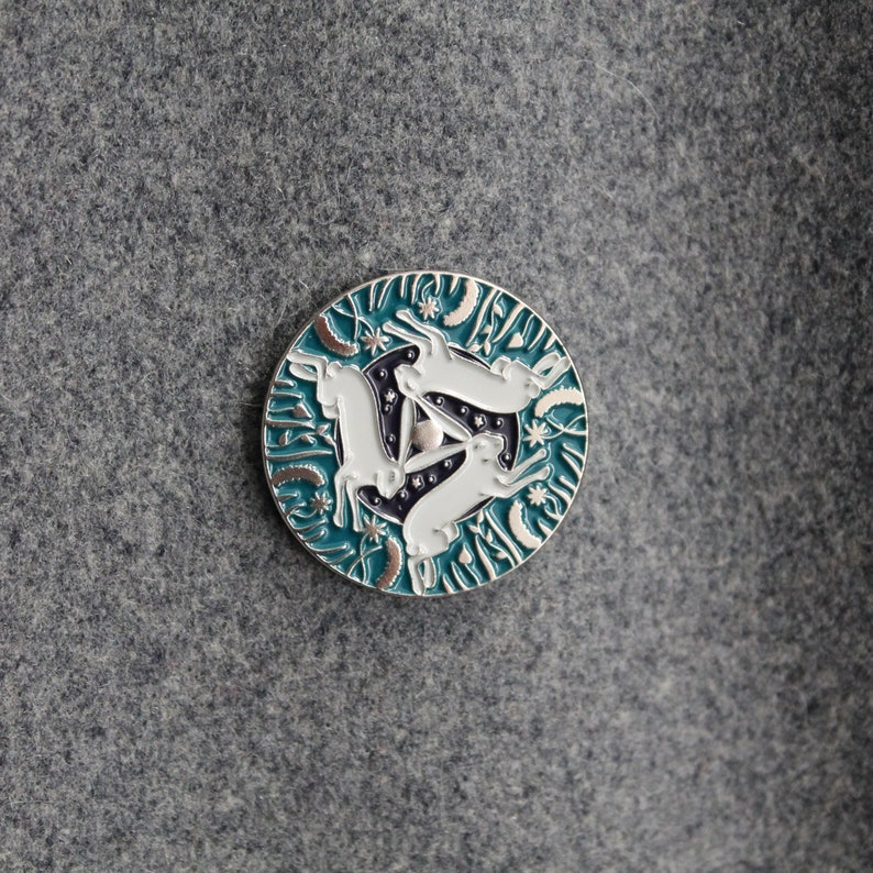 May include: A teal blue enamel pin with a silver border featuring three white rabbits in a circle. The rabbits are in a hopping position and are surrounded by a pattern of stars and leaves.